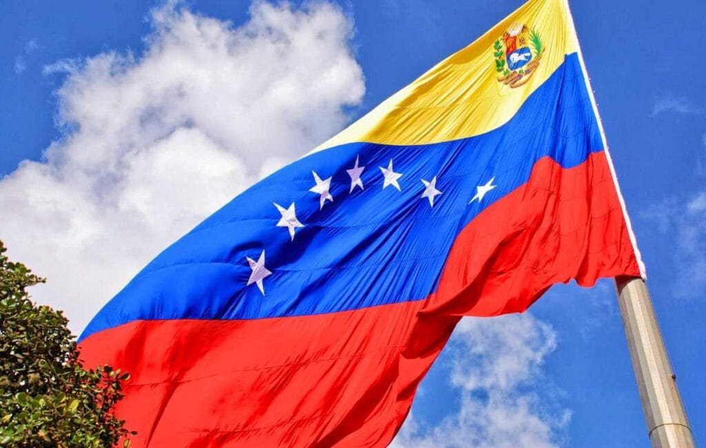 Bandeira da Venezuela amarela azul vermelha com estrelas brancas e brasão ondulando ao vento contra céu azul nublado