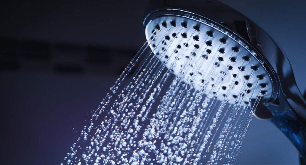 Chuveiro moderno com jato de água caindo em gotículas, em ambiente iluminado por luz azulada, transmitindo sensação de frescor e limpeza. Chuveiro híbrido promete aquecer água com menos energia e reduzir conta de luz em até 70%