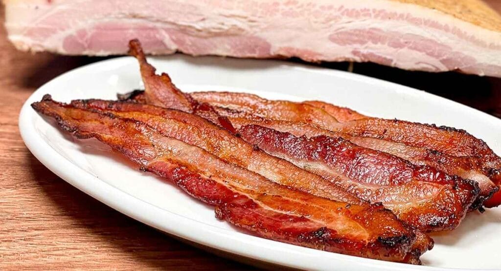 Fatias de bacon crocante dispostas em um prato branco, com um pedaço de carne crua ao fundo, sobre uma mesa de madeira, perfeito para sites de culinária, receitas ou gastronomia. Alimentos cancerígenos