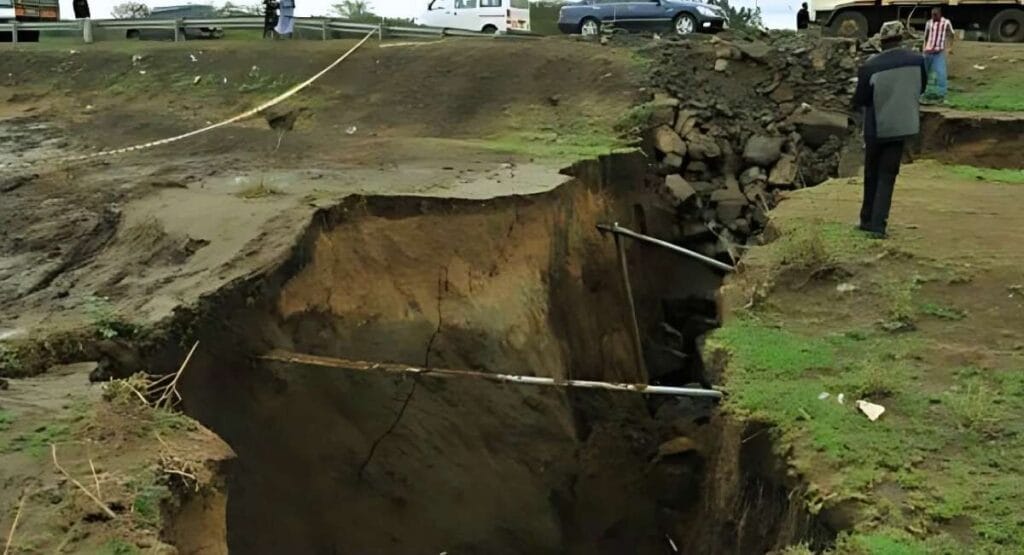 Grande rachadura no solo com tubos expostos, cercada por rochas e pessoas observando perto de estrada.