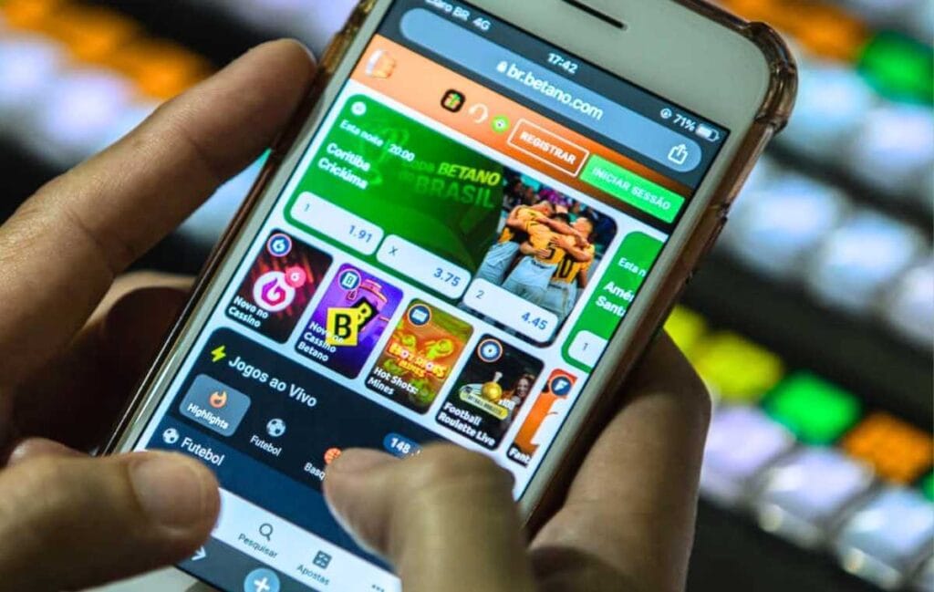 Mão segurando celular exibindo site de apostas Betano com opções de futebol e cassino em fundo desfocado Vício