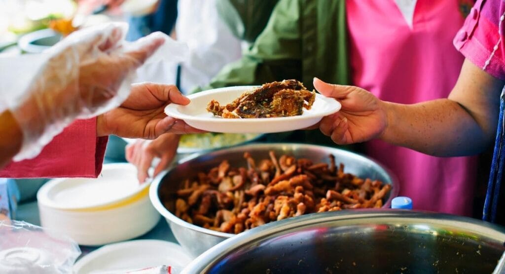 Prato branco com pedaço de frango frito sendo servido por mãos com luvas, próximo a tigela de comida. Fome no mundo