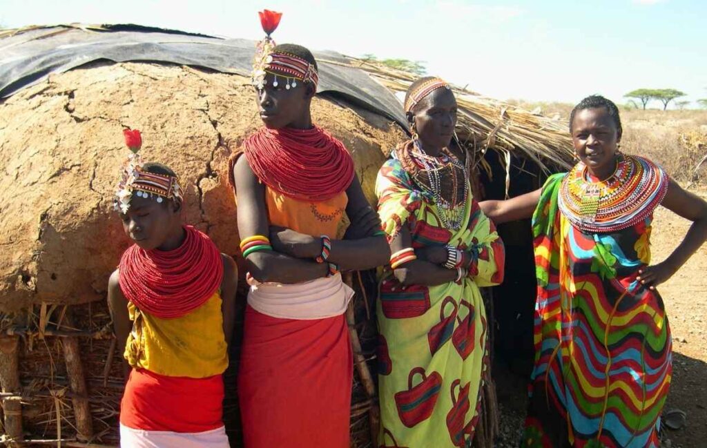 África Quatro mulheres Maasai com colares de contas vermelhos e roupas coloridas posando em frente a cabana de barro