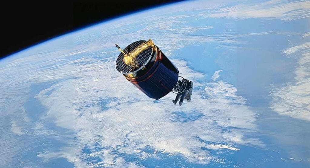 Satélite com antena dourada orbitando a Terra, sobre camadas de nuvens em fundo azul. Astronauta Dale Gardner flutuou no espaço sem cordas para capturar um satélite perdido e o fez com um arpão