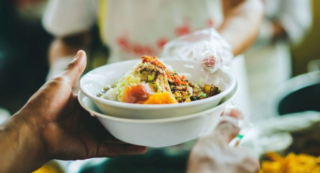 Tigela branca com salada colorida sendo entregue por mãos com luvas, em ambiente de serviço de comida. Fome no mundo está caindo. Fome no mundo