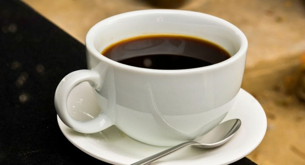 Uma xícara branca de café preto colocada sobre um pires, com uma colher ao lado, sobre uma superfície de madeira com fundo desfocado, ideal para blogs ou sites de culinária e lifestyle. Estudo mostra que quem beber café todos os dias faz bem, tem menos risco de doenças e vive mais