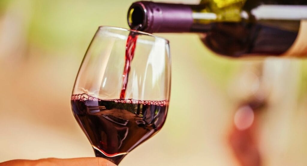 Vinho tinto sendo servido de uma garrafa em uma taça de vidro, com fundo desfocado e tons verdes, ideal para sites de vinícolas, degustações ou lifestyle. Substâncias do vinho podem eliminar bactérias orais
