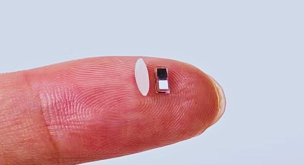 close up da ponta de um dedo, onde estão lado a lado um grão de arroz e um microcomponente eletrônico de tamanho semelhante. Marco-passo