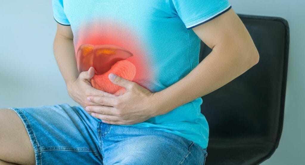 homem com camisa azul e jeans sentado, segurando abdômen com destaque vermelho no fígado, indicando dor ou problema hepático, em ambiente claro hepatite A
