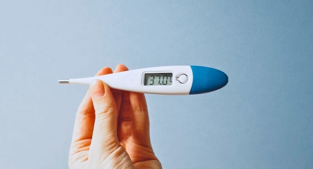 mão com luva segurando termômetro digital exibindo 37.5°C, contra fundo rosa claro, indicando medição de temperatura corporal. febre