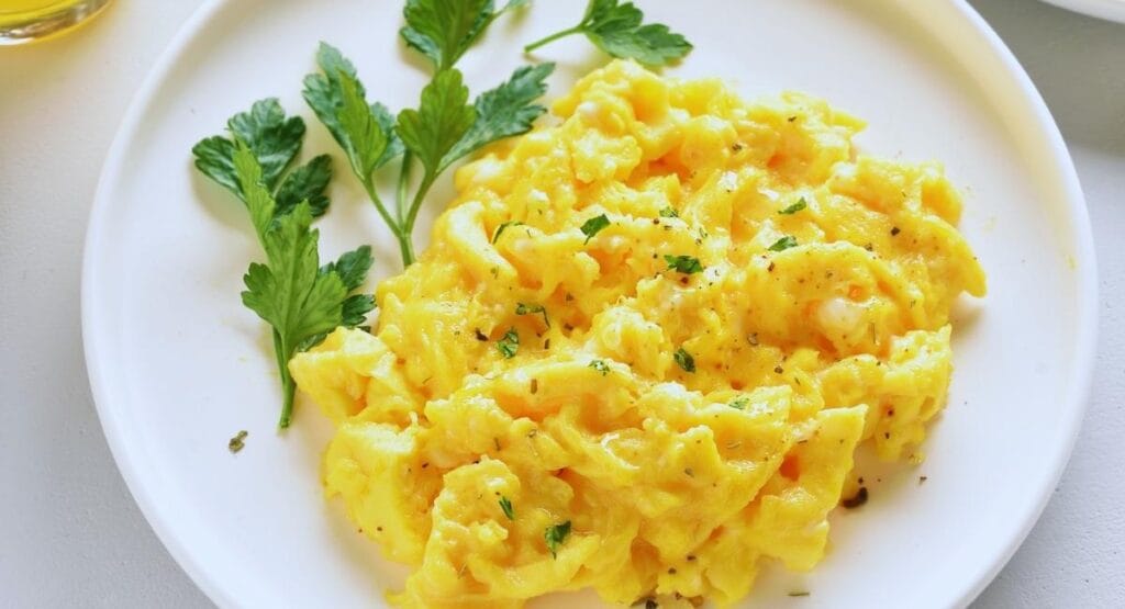 prato branco com ovos mexidos amarelos e fofos, decorados com folhas verdes de salsa, sugerindo uma refeição leve e saudável,