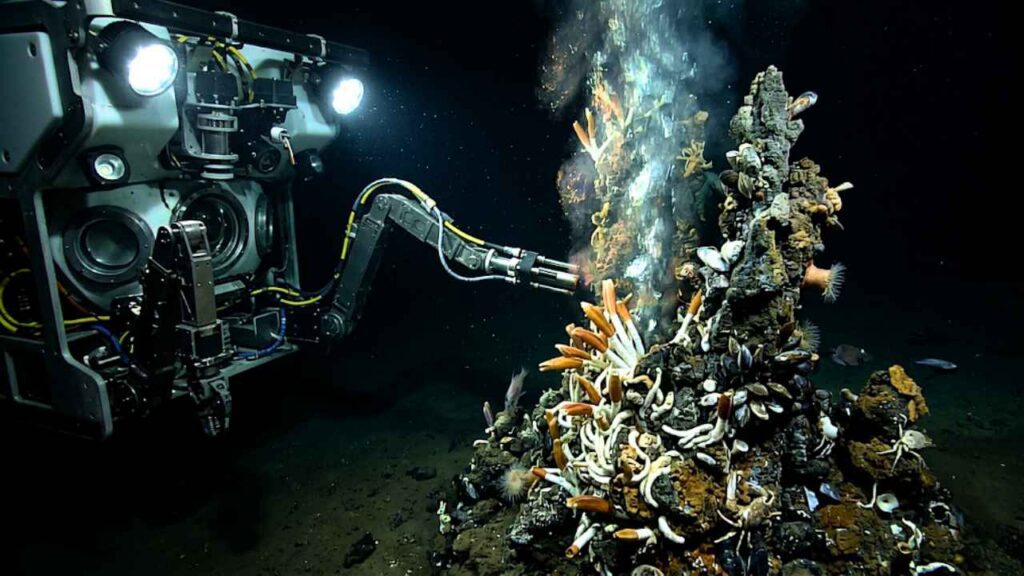 Um Veículo Operado Remotamente (ROV) no fundo do mar estende um braço robótico em direção a uma fonte hidrotermal ativa expelindo fumaça preta. A chaminé mineral ao redor da ventilação está coberta por uma colônia densa de organismos quimiossintéticos, incluindo vermes tubulares de cor laranja brilhante e amontoados de mexilhões do mar profundo, todos iluminados pelas luzes do submersível na escuridão abissal.