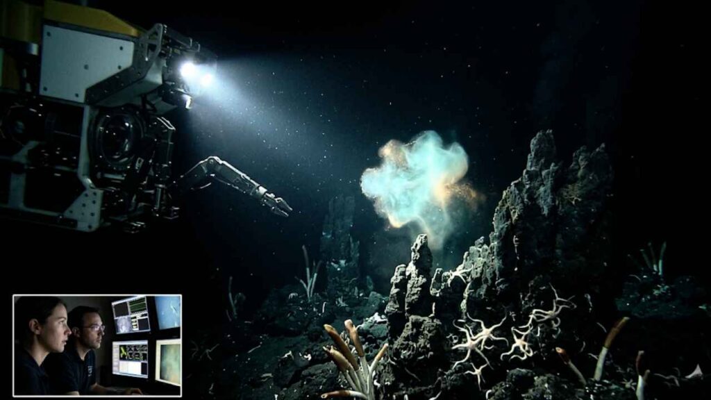 Submarino robótico ilumina uma nuvem brilhante azul e dourada flutuando sobre formações rochosas e corais no fundo do mar escuro, com uma pequena tela no canto mostrando pesquisadores observando a cena.