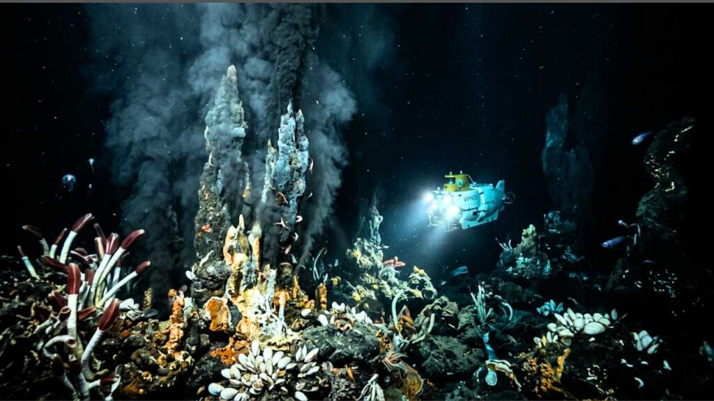 Uma imagem de um submersível operado remotamente (ROV) com o logo WHOI, iluminado por holofotes brilhantes no fundo do mar profundo ao lado de chaminés vulcânicas de ventilação hidrotermal fumegantes, cercadas por animais de águas profundas, incluindo vermes-tubo vermelhos, bivalves e criaturas bioluminosas, flanqueadas por formações rochosas escuras.
