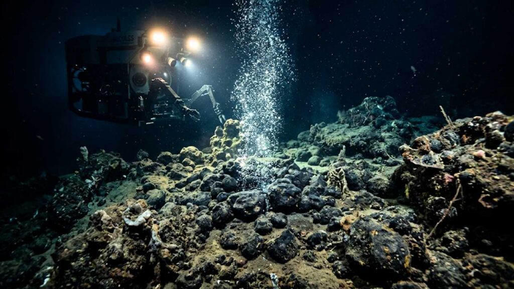Um submersível operado remotamente (ROV Hercules) usa seus holofotes e braços mecânicos para explorar o solo rochoso e escuro do oceano profundo, onde uma coluna de bolhas sobe de uma abertura no fundo do mar.
