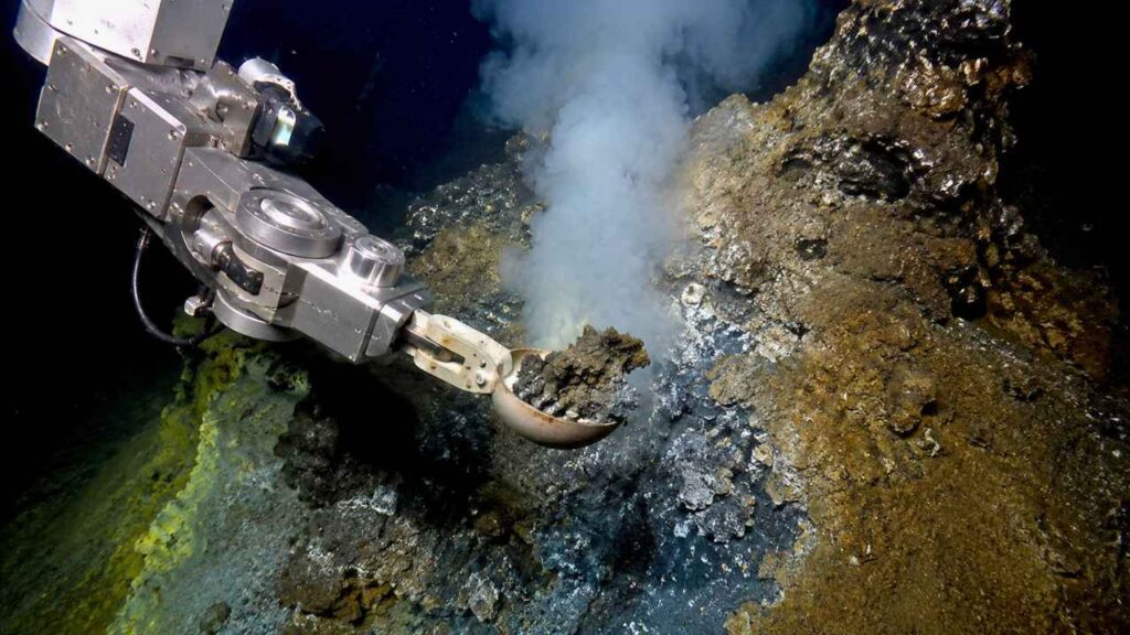Braço robótico explora sedimentos no fundo do oceano a mais de 10 mil metros de profundidade, em uma região escura próxima a fontes hidrotermais onde cientistas estudam microrganismos extremos e os limites da vida na Terra.